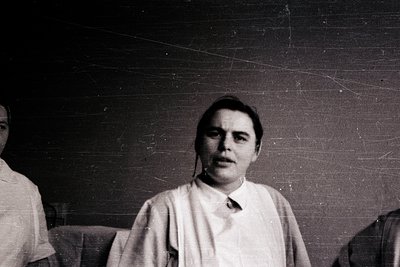 Black-and-white portrait of a man in a high-collared shirt, seated indoors with visible wall cracks. Likely mid-20th century ...