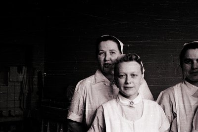 Three nurses in early 20th-century uniforms pose indoors, likely a hospital or clinic. White starched collars, caps, and long...