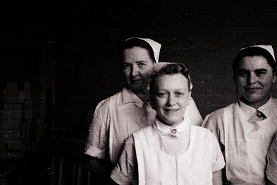 Three nurses in early 20th-century uniforms pose indoors, likely a hospital or clinic. White starched caps, collared blouses,...
