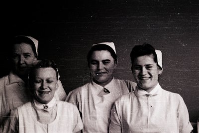 Four nurses in early 20th-century uniforms pose indoors, likely a clinic or hospital. White caps, collared blouses, and dark ...