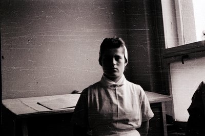Vintage black-and-white portrait of a young individual in a high-necked blouse, seated indoors near a wooden desk with a wind...