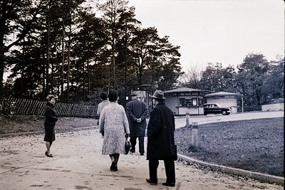 Mid-20th century street scene featuring four pedestrians in vintage attire—men in suits, women in long coats—walking along a ...