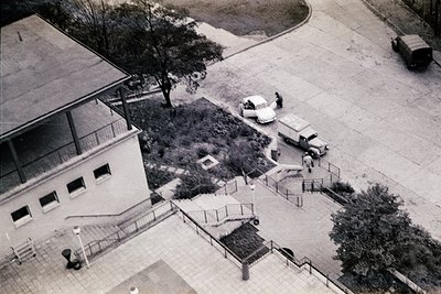 Aerial view of a mid-20th-century courtyard with a two-story building featuring flat roof and rectangular windows. Two vintag...