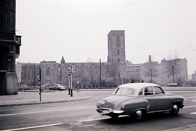 Vintage black-and-white street scene featuring a mid-century classic car driving past a grand, Gothic-style church with a tal...