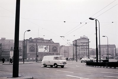 Black-and-white urban street scene featuring mid-20th century architecture. A vintage van drives past a billboard advertising...