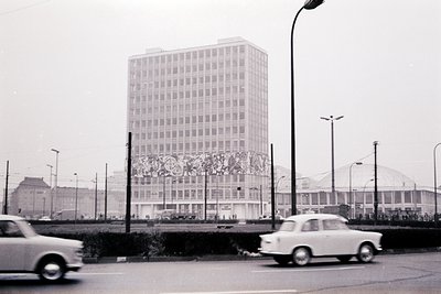 1960s-era high-rise with Brutalist concrete facade, featuring geometric mural. Circular stadium with dome roof in background....
