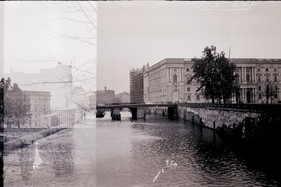 Vintage black-and-white urban riverside scene featuring a stone bridge spanning a calm waterway. Symmetrical buildings with c...