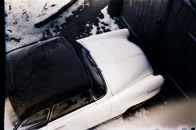 Vintage sedan parked in a snow-covered alley, likely mid-20th century. Dark sedan (left) contrasts with faded white sedan (ri...