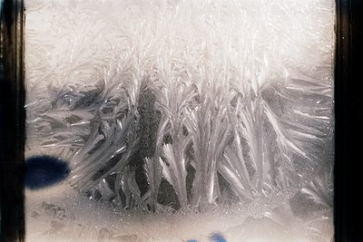 Close-up of intricate frost patterns on a windowpane, resembling delicate lacework. The crystalline formations suggest cold w...