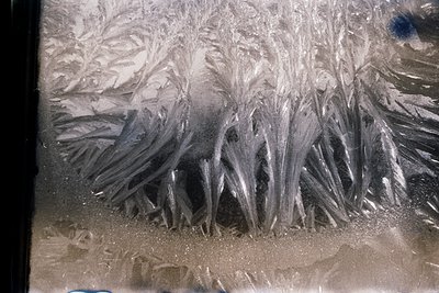 Close-up of intricate frost patterns on a windowpane, resembling delicate ice crystals or abstract art. The texture suggests ...