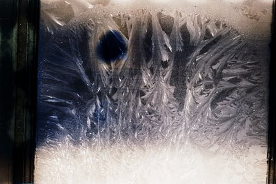 Close-up of intricate **frosted window patterns** on a pane, featuring symmetrical ice crystal formations resembling **alpine...