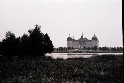 Baroque-style castle with twin domed towers and central dome, set beside a reflective moat. Lush greenery frames the foregrou...