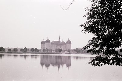Neoclassical palace reflected in a serene lake, framed by foliage. Symmetrical dome-topped wings and central tower suggest 18...