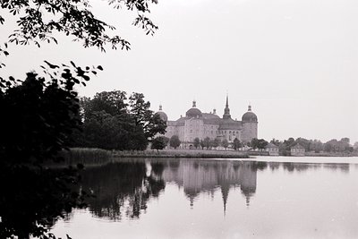 Historic Baroque-style castle reflected in calm waters, surrounded by dense greenery. Likely 18th–19th century European archi...