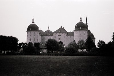 Symmetrical Baroque-style castle with two prominent domed towers flanking a central entrance. Lush greenery surrounds the sto...