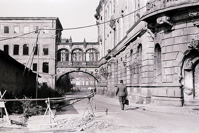 Historic black-and-white street scene featuring a man walking near a grand stone archway with ornate carvings. The archway le...