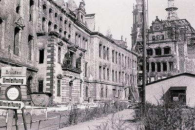 War-damaged European cityscape, likely post-WWII. Prominent multi-story buildings with ornate facades show severe structural ...