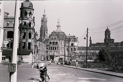 Mid-20th century urban street scene featuring historic European architecture. Prominent domed and spired buildings with ornat...