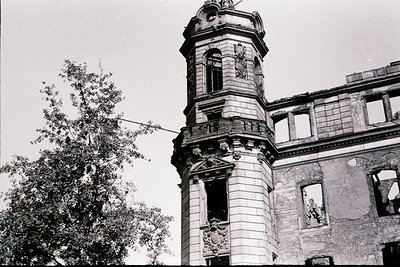 Dilapidated neoclassical tower with ornate stonework and arched openings, flanked by crumbling walls. Decorative reliefs and ...