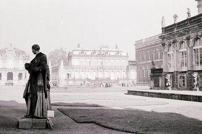 Neoclassical courtyard featuring a prominent statue of a robed figure in profile, likely a historical or royal portrait. Symm...