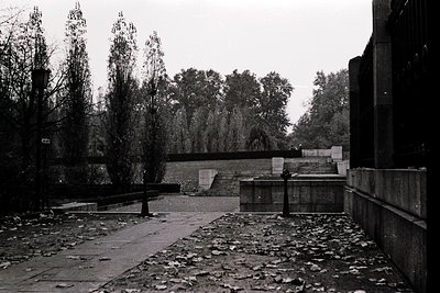 A stark, monochrome courtyard featuring brutalist concrete architecture. Low walls, planters, and a central pathway flanked b...