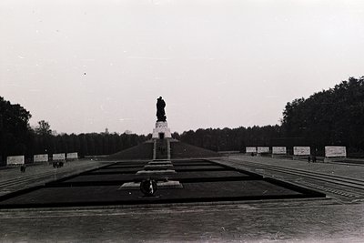 Monumental statue atop tiered stone platform in a wide, symmetrical plaza flanked by banners and trees. Likely a Soviet-era m...