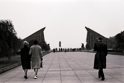 Monumental Soviet-style plaza with symmetrical concrete pyramids flanking a central statue. Mid-20th century attire suggests ...