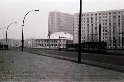 Black-and-white urban street scene featuring Soviet-era Brutalist architecture. Prominent curved-roof building with geometric...
