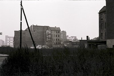 Black-and-white urban ruin featuring a partially collapsed brick building, likely post-war or mid-20th century. Foreground sh...