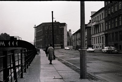 Black-and-white street scene featuring a pedestrian bridge with a single person walking toward the camera. Industrial-era bui...