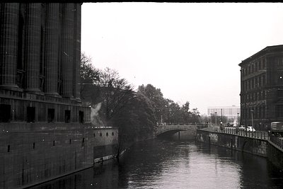 Black-and-white urban canal scene featuring industrial-era architecture. Prominent brick buildings with large windows flank a...