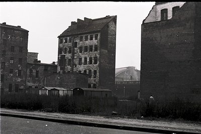 Mid-20th century urban decay: multi-story brick buildings with exposed wiring, missing windows, and crumbling facades. Overgr...