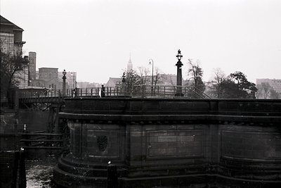 Black-and-white urban scene featuring a historic bridge with ornate lampposts and a central monument. The structure’s curved ...
