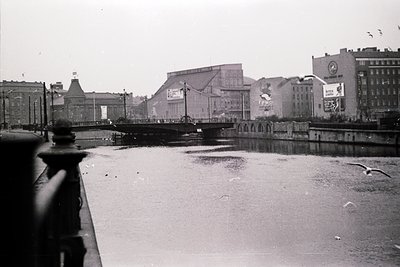Black-and-white urban scene featuring a bridge spanning a calm river, flanked by mid-20th-century Soviet-era architecture. Pr...