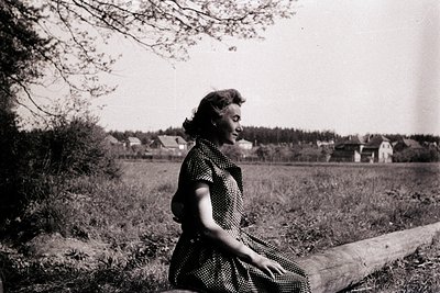 Black-and-white portrait of a woman seated on a fallen log in a rural landscape, mid-20th century. She wears a polka-dot dres...