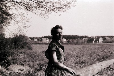 Mid-century woman in a polka-dot dress poses outdoors beside a rustic wooden fence, set against a rural landscape with scatte...