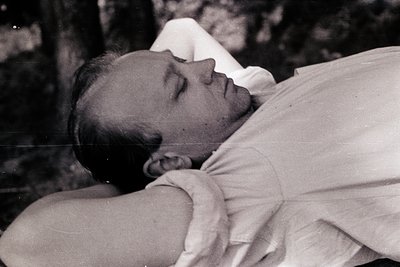 Black-and-white portrait of a man reclining outdoors, likely mid-20th century. He wears a loose, high-collared shirt and rest...