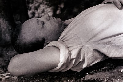 Black-and-white portrait of a reclining man in a loose, high-collared shirt, resting on a rough stone surface. The compositio...