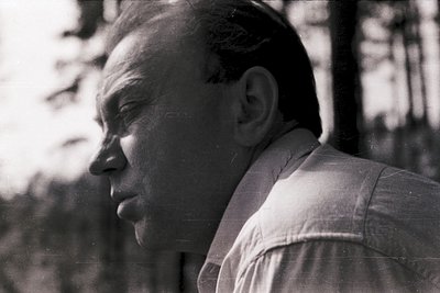 Portrait of a man in mid-speech, outdoors in a wooded area. Lighting suggests midday sun filtering through trees. Clothing in...