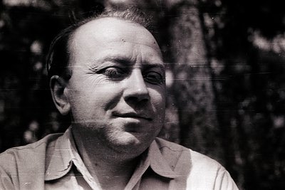 Mid-century black-and-white portrait of a man in a light-colored button-up shirt, set against blurred foliage. His expression...
