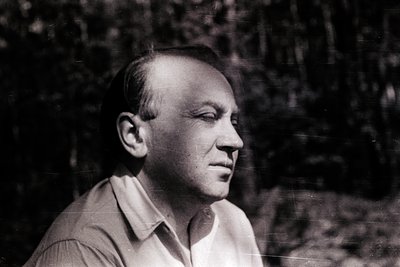 Black-and-white portrait of a man in mid-profile, set against a blurred forest backdrop. Lighting suggests midday outdoor set...
