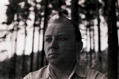 Black-and-white portrait of a man in a forest setting, likely mid-20th century. His short hair and neutral expression suggest...