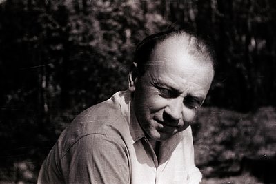 Mid-century black-and-white portrait of a man in a light button-down shirt, set against blurred greenery. His thoughtful expr...