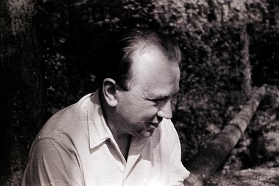 Mid-century black-and-white portrait of a man in a light button-down shirt, seated outdoors among dense foliage. His thoughtf...