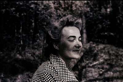 Portrait of a woman in mid-laugh, set against blurred forest foliage. She wears a polka-dot blouse with a high, voluminous ha...