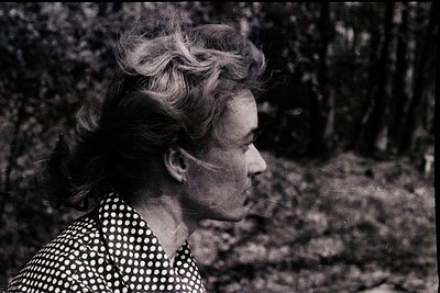 Black-and-white portrait of a woman in mid-profile, set against a blurred forest backdrop. She wears a polka-dot blouse and h...