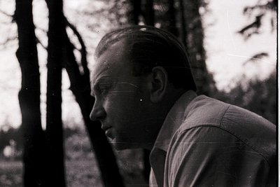 Black-and-white portrait of a man in profile, outdoors among blurred trees. Mid-century hairstyle and button-up shirt suggest...