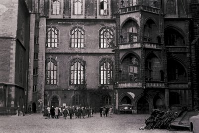 Gothic-style courtyard with stone façade featuring pointed arches and large rectangular windows. Group of people in 1940s-era...