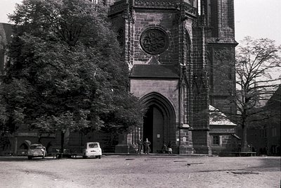Gothic-style stone church facade with pointed arches, intricate carvings, and a central rose window. Two parked vintage cars ...