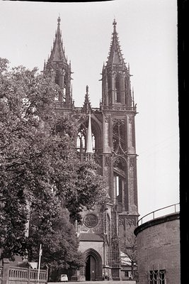 Gothic cathedral façade with twin spires and intricate tracery, framed by mature trees. Circular clock near the entrance and ...